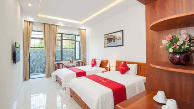 Bright Hoi An hotel room with two beds, red accents, a window, wooden furniture, and flowers on a shelf.
