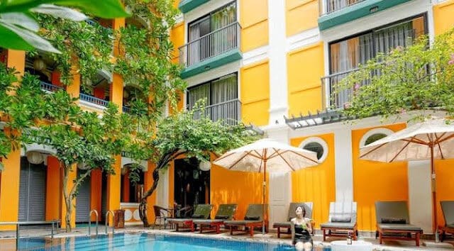 A woman sits by a pool at a bright yellow Hoi An hotel, surrounded by balconies and poolside lounge chairs.