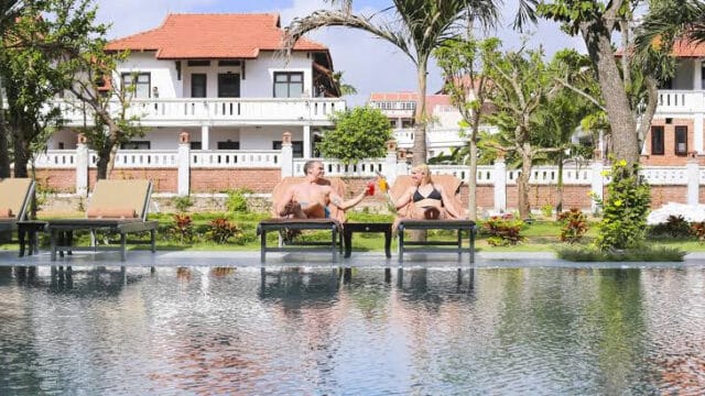 Two people relax on loungers by a pool in Hoi An, sipping drinks with villas and palm trees in the background.