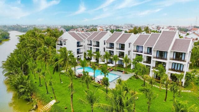Aerial view of a modern Hoi An riverside resort with a pool, palm trees, and manicured green lawns.