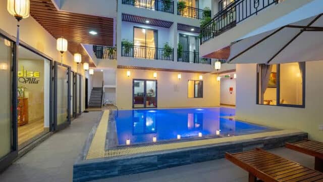 A well-lit indoor swimming pool area with lounge chairs, balconies above, and Hoi An-inspired decor.