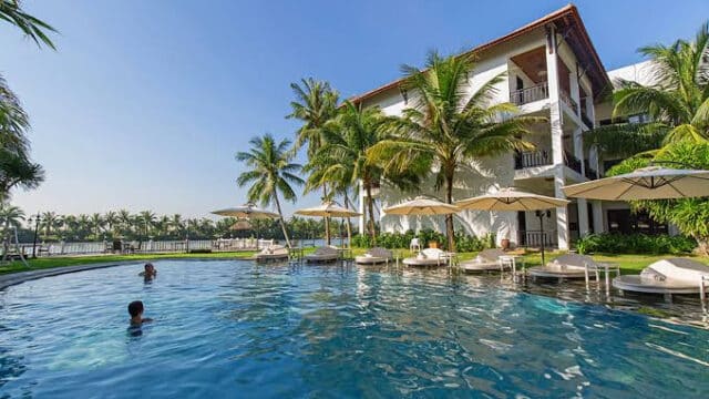 Resort pool with sun loungers, palm trees, and people swimming beside a white building in Hoi An under a clear sky.