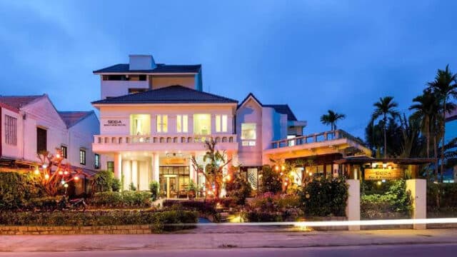 Modern, well-lit Hoi An house with large windows and balconies, surrounded by plants and trees, at dusk.