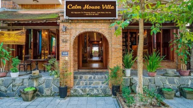 Brick-walled entrance of Calm House Villa in Hoi An with plants and a yellow sign above the doorway on a sunny day.