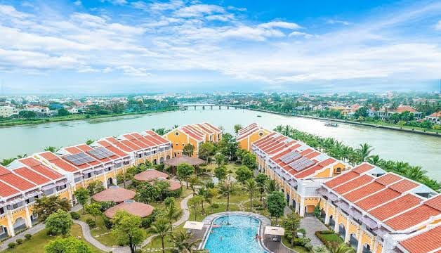 Aerial view of a riverside Hoi An resort with red-roofed buildings, palm trees, and a large swimming pool.