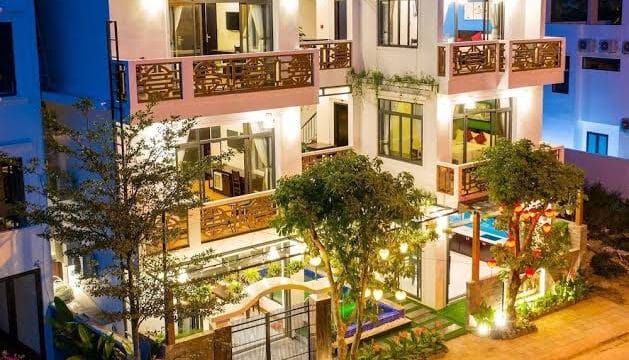 Three-story modern house in Hoi An with balconies, large windows, outdoor lights, and trees in the front at night.