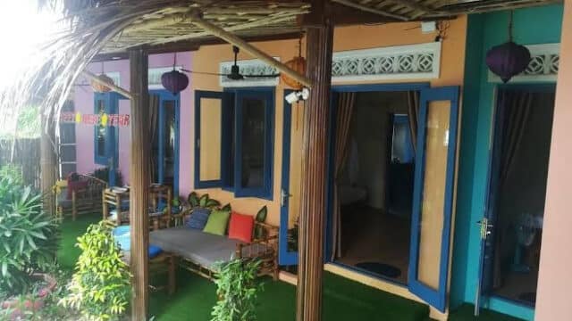 Colorful Hoi An bungalow porch with bamboo pillars, outdoor seating, and open doors beside green plants.