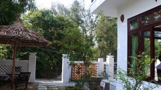 A cozy garden patio in Hoi An with a thatched umbrella, lush plants, and a white modern house with large windows.