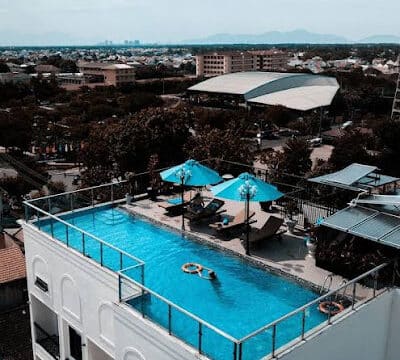 Rooftop infinity pool with blue umbrellas, lounge chairs, and a stunning Hoi An cityscape view in the background.
