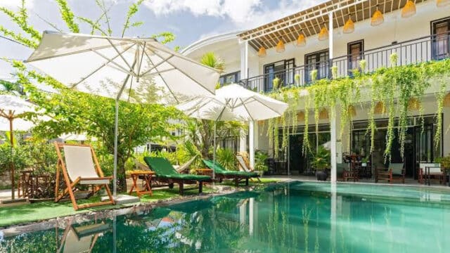 Outdoor pool with lounge chairs and umbrellas beside a modern Hoi An building featuring hanging plants and a balcony.