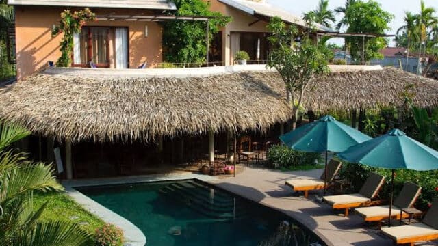 A tropical villa in Hoi An with a thatched roof, pool, sun loungers, and green umbrellas surrounded by lush plants.