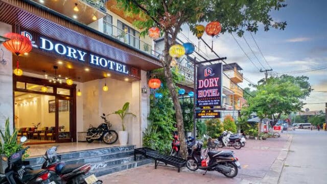 Dory Hotel entrance in Hoi An with motorbikes parked outside, colorful lanterns, and a tree-lined street.