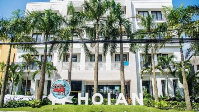 White building with palm trees and large HOIAN letters on the lawn, set under Hoi An’s clear blue sky.