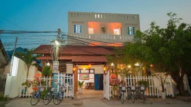 Two-story Hoi An guesthouse with lights on, white fence, bicycles outside, and trees on each side at dusk.