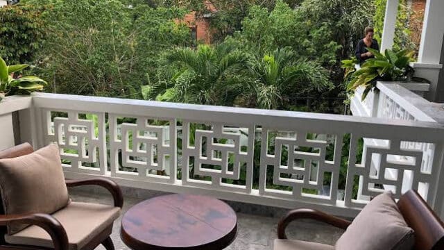 Two cushioned chairs and a round table on a Hoi An balcony overlooking lush green trees and plants.
