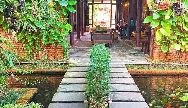 Stone path over a koi pond leads to a cozy, Hoi An-inspired café with brick walls, wooden furniture, and lush plants.