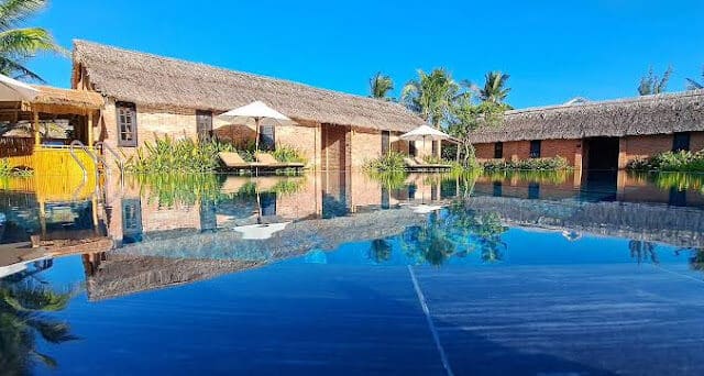 Thatched-roof cottages and umbrellas reflected in a calm, blue Hoi An swimming pool on a sunny day.