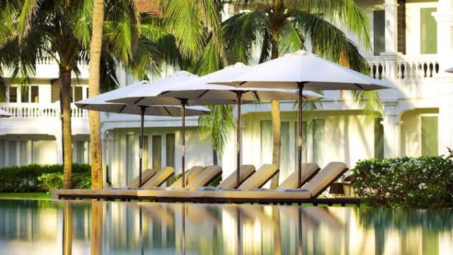 Lounge chairs and umbrellas by a pool in Hoi An, with palm trees and a white resort building in the background.