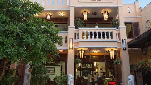 The front of a peach-colored Hoi An hotel with balconies, lanterns, and greenery, with people gathered at the entrance.