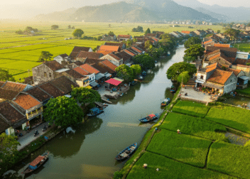 An aerial view of a traditional Vietnamese riverside village with terracotta-roofed houses, lush green rice paddies, narrow waterways with boats, and mountainous backdrop in the distance.