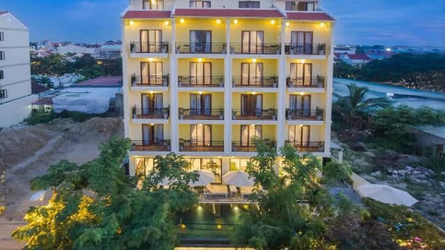 A multi-story yellow hotel building with balconies, pool, and Hoi An greenery illuminated at dusk.