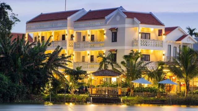 A well-lit, three-story riverside hotel in Hoi An with balconies, palm trees, and outdoor seating at dusk.