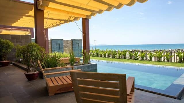 Sunny patio in Hoi An with wooden chairs, pool, green plants, ocean view, all under a yellow canopy.