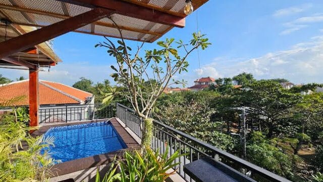 A rooftop balcony in Hoi An with a small pool, lush plants, and views of trees and red-roofed houses beneath a blue sky.