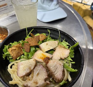 A bowl of Hoi An noodles with pork, greens, and crispy toppings sits beside a drink and a light blue cap on the table.