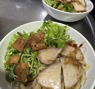 Two bowls of noodles topped with sliced pork, fresh greens, and crispy toppings on a metal table in charming Hoi An.