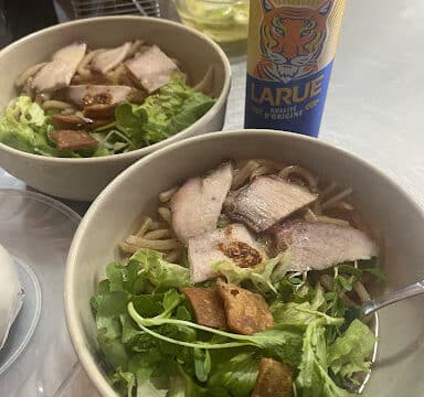 Two bowls of Hoi An noodle soup with greens and pork, next to a can of Larue beer on a table.