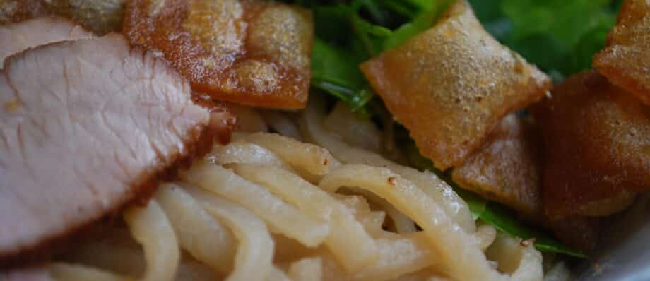 Close-up of a bowl of cao lau with noodles, pork, herbs and croutons