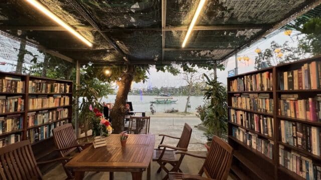 Outdoor library with wooden tables and chairs, bookshelves, and a river view in the background at sunset.