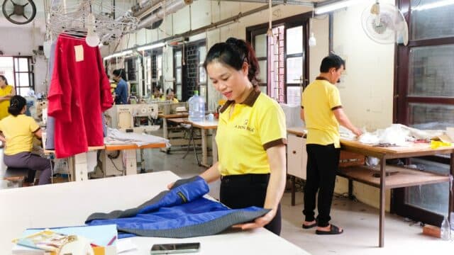 Woman folding fabric in a brightly lit garment factory with other workers in yellow shirts in the background.