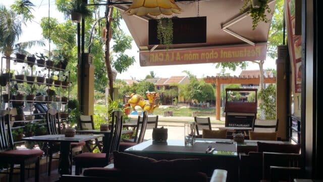 Outdoor view from inside À La Carte Café with cozy wooden chairs, plants, and sunlight streaming through the entrance.