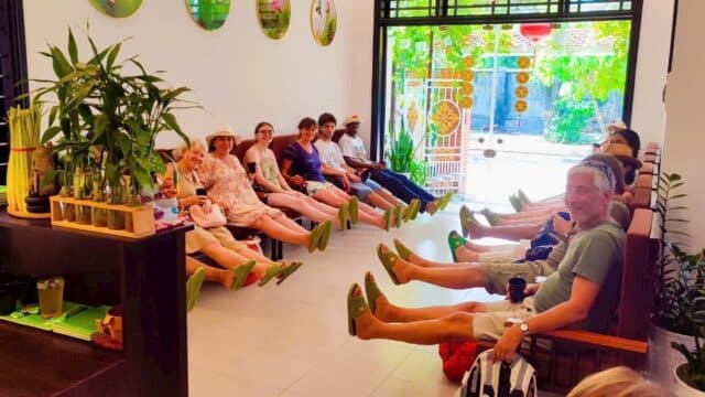 A group of people sit indoors at Aly Garden Spa, wearing green foot coverings and smiling, with plants decorating the space.