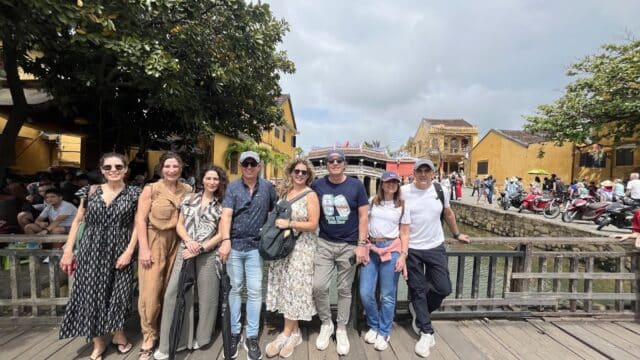 Eight people smile together on a wooden bridge in a historic town, enjoying their day with Amigos Tours among charming yellow buildings.