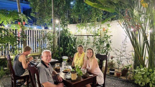 Four people dining outdoors at a Hoi An garden restaurant at night, surrounded by lush plants and glowing string lights.