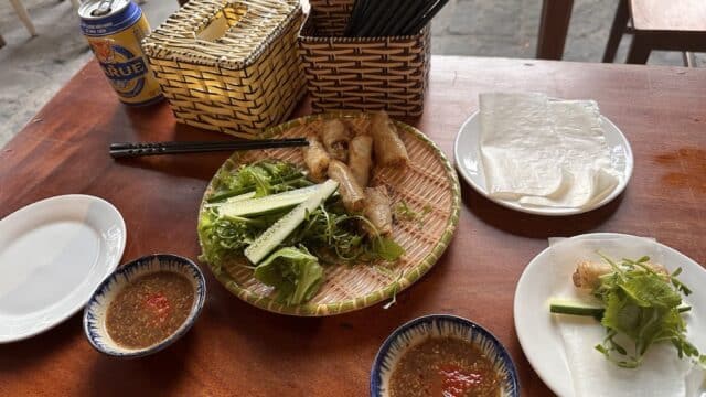A Vietnamese meal at An Hy Vegetarian Restaurant with spring rolls, greens, rice paper, dipping sauce, and a beer on a wooden table.