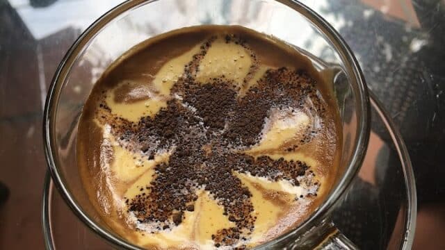 A glass cup of coffee with a flower-shaped cocoa powder design, reflecting Hoi An’s charm on a glass table.