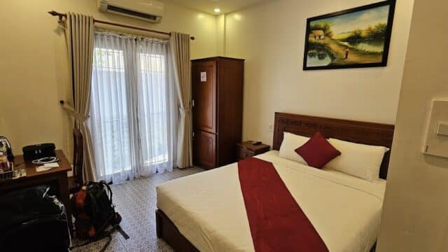 Cozy Ancient Hoi An hotel room with a double bed, red runner, wardrobe, desk, and sunlight through curtained glass doors.
