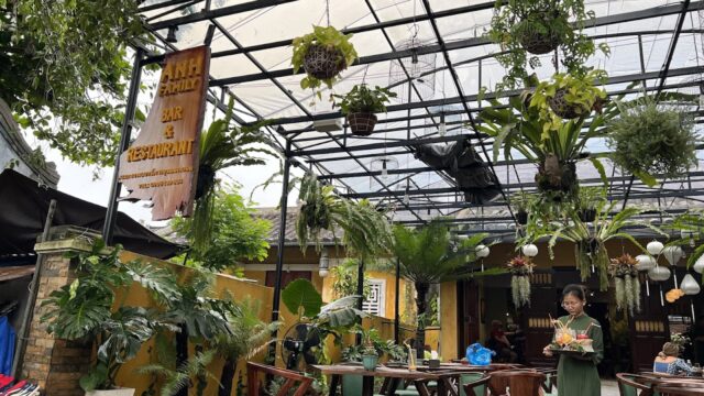 Covered outdoor restaurant, Anh Family Bar, with many hanging plants, wooden tables, and a server carrying drinks.