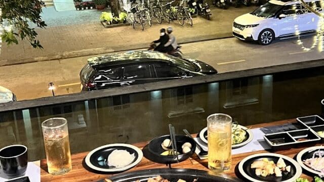 Korean BBQ grill with meat and sides on a table at Apkujeong BBQ, overlooking a lively street with lights and parked vehicles.