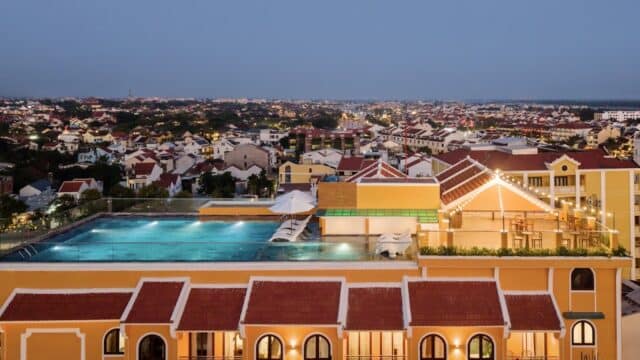 Rooftop pool at Aria Boutique Hotel on a yellow-orange building, overlooking a cityscape at dusk with sparkling lights and houses.