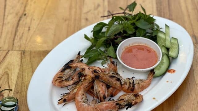 Grilled shrimp with dipping sauce, greens, and cucumber slices on a white plate—a taste of Hoi An—served on a wooden table.