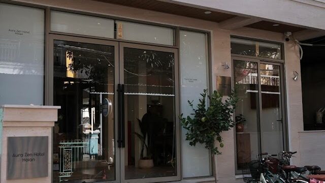 Modern building entrance of Aura Zen Hotel with glass doors, potted plant, parked bicycles, and a red scooter outside.