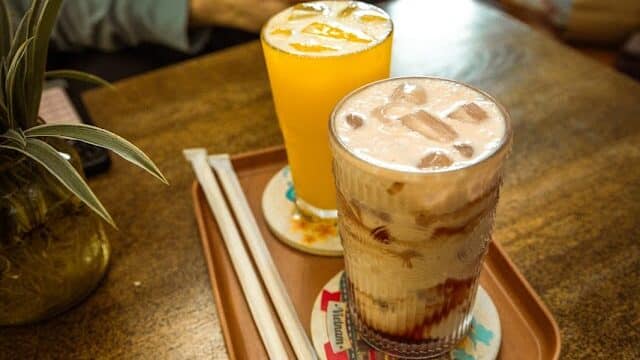 Two drinks—one orange, one iced coffee—on a tray at Avocado Cafe; straws ready, person seated behind at a wooden table.