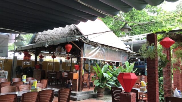 Outdoor restaurant with brown chairs, red lanterns, and green plants under partial awnings and a thatched roof.