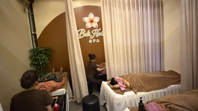 Three people unwind with massages in a cozy Bali Herbal Spa room adorned with cream curtains and a spa sign on the wall.