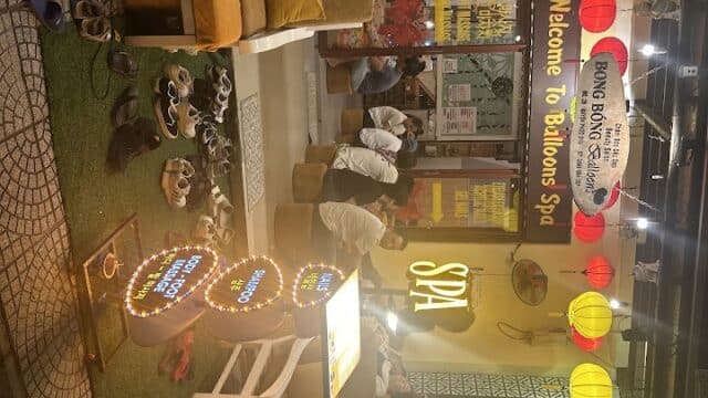 People seated on benches outside Balloons Spa, with colorful lanterns and many shoes by the brightly lit entrance.